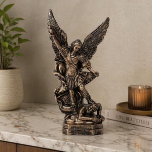 Op de afbeelding: Bronskleurig beeld van een gevleugelde figuur met een zwaard, staand over een verslagen figuur. Het beeld heeft ingewikkelde details en staat op een rechthoekige basis. Een decoratief item.