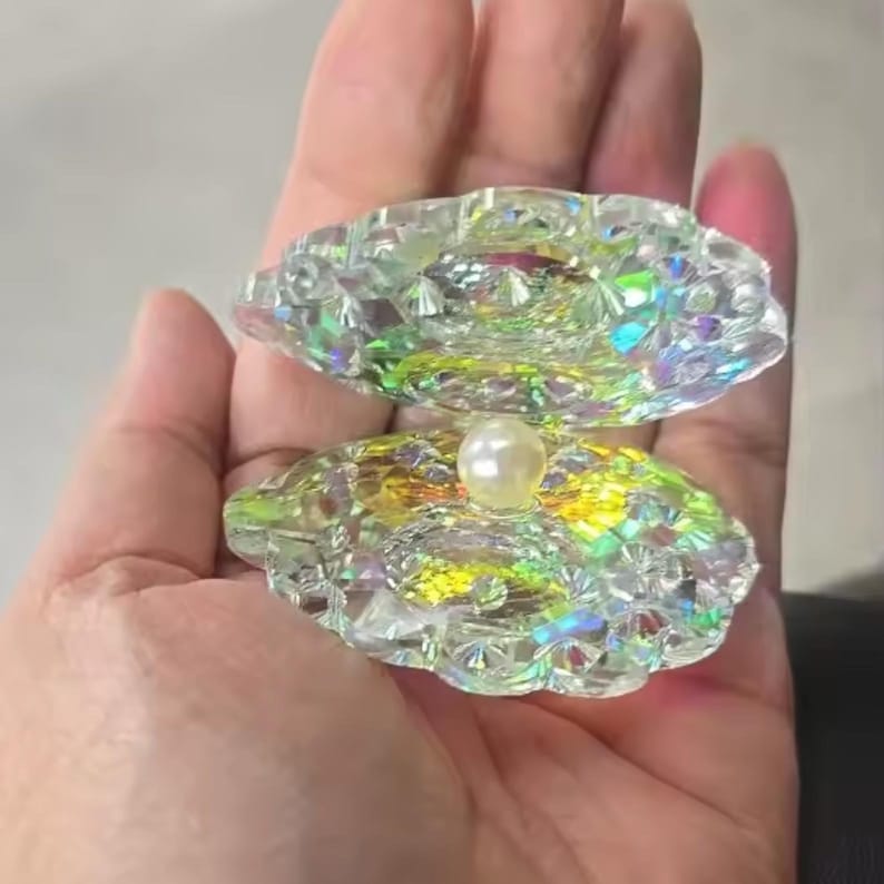 Pode incluir: Uma concha decorativa aberta feita de vidro transparente e facetado. A concha tem reflexos iridescentes de arco-&iacute;ris e cont&eacute;m uma &uacute;nica p&eacute;rola branca e redonda. A concha &eacute; segurada na m&atilde;o de uma pessoa.