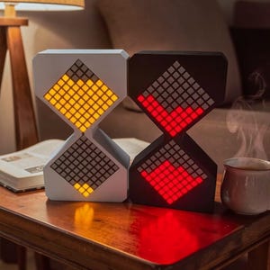 Puede incluir: Dos relojes digitales con forma de reloj de arena. Uno es blanco con luces amarillas, el otro negro con luces rojas. Están sobre una superficie de madera, con un libro y una taza de té cerca. Los relojes tienen un diseño pixelado.