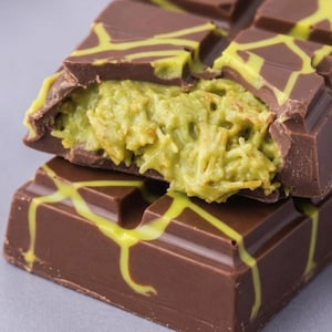 May include: A close-up of a chocolate bar with a pistachio filling. The dark chocolate is decorated with yellow-green lines. The bar is broken open, revealing the green filling. The image is set against a gray background.