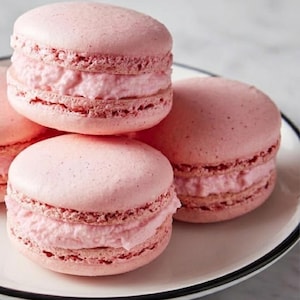 Può includere: Primo piano di macarons rosa su un piatto bianco con bordo nero. Ogni macaron ha un guscio rosa chiaro e un ripieno cremoso rosa. I macarons sono impilati e disposti per mostrare la loro consistenza e il loro colore.