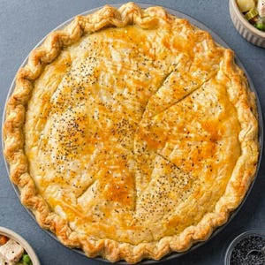 May include: A golden-brown, freshly baked pie with a flaky crust, cut into wedges. The pie is sprinkled with black seeds and sits on a round, gray plate. The crust has a decorative, crimped edge. A culinary delight.