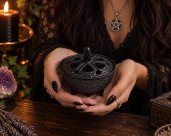 May include: A small, round, black cast iron cauldron with three short legs and a removable lid featuring a cut-out star pattern and a central handle. The exterior surface has a textured, matte finish, and the lid is designed to allow smoke to escape.