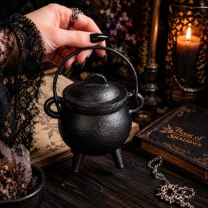 May include: A small, round, black cast iron cauldron with three short legs, a matching lid with a central handle, and a curved metal swing handle attached to two side loops. The exterior surface has a textured, matte finish.