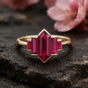 以下が含まれることがあります： 中央に細長いルビー色の宝石をあしらったゴールドリング。両側に2つの小さな宝石が配置されています。リングは幾何学的なデザインで、暗く質感のある表面に置かれています。