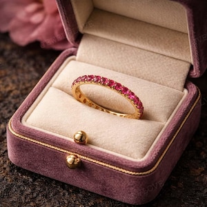 May include: A gold ring with a band of small, vibrant red gemstones, presented in a purple velvet jewelry box. The ring is centered in the box, showcasing its design. The box has a gold trim and a soft, plush interior.