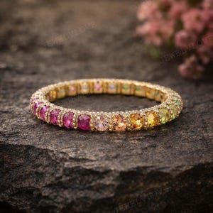 Puede incluir: Un anillo de oro con una banda de piedras preciosas de colores. Las piedras preciosas cambian de color de rosa a amarillo y verde. El anillo está sobre una superficie oscura y texturizada.