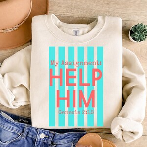 Könnte beinhalten: Cremefarbenes Sweatshirt mit dem Text "My Assignment: HELP HIM Genesis 2:18" in Koralle und Türkis. Der Text ist mittig auf der Vorderseite platziert, mit türkisfarbenen vertikalen Streifen hinter den Worten. Das Sweatshirt ist gefaltet.
