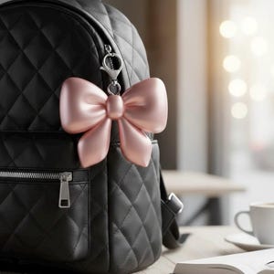 Puede incluir: Una mochila negra acolchada con cremallera plateada y un llavero en forma de lazo rosa adjunto. El lazo es de color rosa pastel suave y tiene un acabado brillante y liso. La mochila está sobre una mesa de madera.