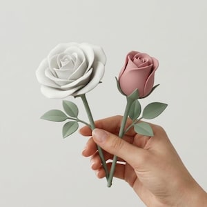 Può includere: Una mano tiene due rose artificiali: una bianca, completamente aperta, e un bocciolo rosa. Entrambe hanno steli e foglie verdi. Lo sfondo è semplice. Le rose sono probabilmente fatte di un materiale morbido.