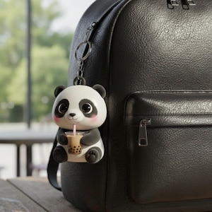 May include: A black and white panda bear keychain with a boba drink, attached to a black backpack. The panda has large eyes and is holding a cup with a pink straw. The backpack is made of textured material.