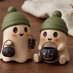 Könnte beinhalten: Zwei cremefarbene Geisterfiguren mit olivgrünen Strickmützen mit Bommeln. Jeder Geist hat ein lächelndes Gesicht mit rosigen Wangen und hält einen kleinen schwarzen Kürbiseimer. Ein Geist hält zusätzlich einen schwarzen Becher mit orangefarbenen Punkten.