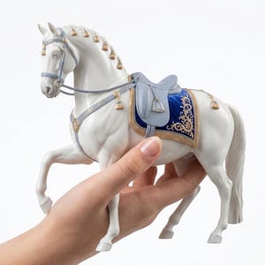 May include: A white horse figurine with a blue saddle and gold trim. The horse has a detailed bridle and decorative accents. The figurine is held in a hand, showcasing its size and design. The horse is posed with one leg raised.