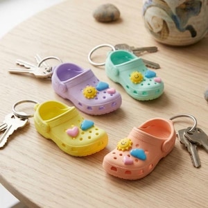 May include: Four miniature, colorful clogs keychains in yellow, purple, mint green, and peach. Each clog features a smiling sun, heart, and cloud embellishments. Keychains are on a light wood surface with keys and a decorative ceramic cup in the background.
