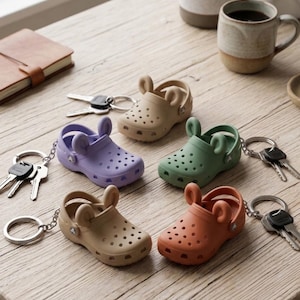 May include: A collection of miniature Crocs keychains in various colors: lavender, green, orange, and two shades of tan. Each keychain features a pair of tiny shoes with bunny ear accents. The keychains are arranged on a wooden surface, with keys and a notebook nearby.