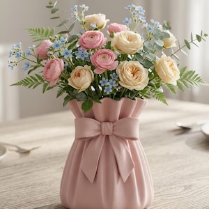 Puede incluir: Un jarrón de cerámica rosa con un lazo, lleno de un ramo de flores rosas, crema y azules. El arreglo incluye rosas, pequeñas flores azules y vegetación, creando una exhibición suave y elegante. El jarrón tiene una parte superior con volantes.