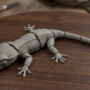 May include: A gray, segmented gecko figurine with articulated limbs, lying on a dark wooden surface. The gecko has a detailed, textured appearance, with visible stitching along its back. The tail is long and slender. A knife is visible in the background.