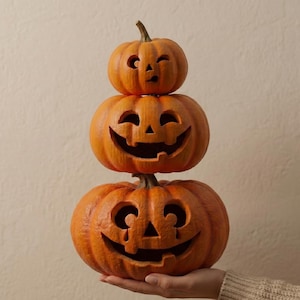 May include: A stack of three carved orange pumpkins with smiling faces, perfect for Halloween decor. The pumpkins are stacked on top of each other, with the smallest pumpkin on top and the largest on the bottom. The pumpkins are lit from within, creating a spooky glow.