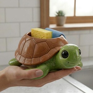 May include: A ceramic turtle-shaped sponge holder. The turtle has a brown shell and a green head and legs. The holder contains a yellow sponge and a blue cloth. The turtle is held in a hand.