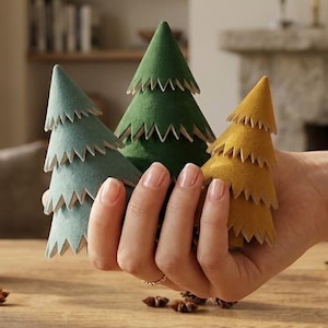 May include: Three small, cone-shaped decorative trees made of felt in muted shades of sage green, forest green, and mustard yellow. Each tree features a layered, jagged-edge design that mimics pine needles, held together in a hand.