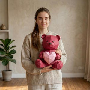 Könnte beinhalten: Ein bordeauxfarbener Papierbär mit geometrischem Design und einem rosa Herz. Der Bär hat eine schwarze Nase und ist ein dekoratives Objekt. Der Bär ist wahrscheinlich ein Geschenk oder ein Einrichtungsgegenstand mit moderner, polygonaler Ästhetik.