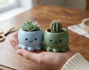 May include: Two small, octopus-shaped planters, one light blue and one green, each holding a succulent. The blue planter has a smiling face, while the green one has a frowning face. The planters are held in a person's hand.