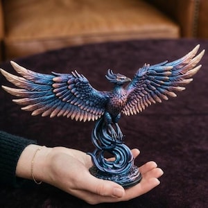 May include: A detailed figurine of a phoenix with outstretched wings. The sculpture features a blend of blue, purple, and gold hues. The phoenix is perched atop a swirling base, showcasing intricate feather details and a regal pose.