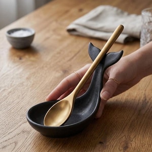 May include: A black ceramic spoon rest shaped like a whale tail, holding a gold-coloured spoon. The spoon rest has a small bowl for the spoon and a curved tail design. The spoon is made of metal and has a long handle.