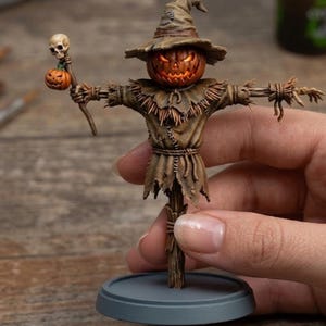 May include: A detailed miniature Halloween scarecrow figurine. The scarecrow has a carved pumpkin head with glowing eyes and mouth, a straw body, and a pointed hat. It holds a staff with a skull and a small pumpkin. The figurine is brown and orange.