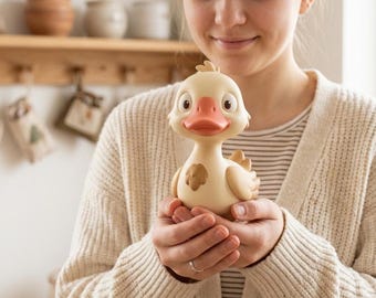 Puede incluir: Un juguete de pato de goma de color crema con un acabado mate suave, un pico prominente de color rosa coral, ojos grandes y oscuros y una pequeña mancha marrón en el pecho. El pato tiene una cabeza redondeada, cuello corto y alas estilizadas.