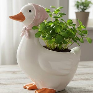 May include: A white ceramic goose planter with an orange beak and feet, wearing a pink bonnet. The planter is filled with green leafy plants. A charming decorative piece for indoor or outdoor use.