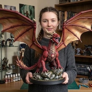 May include: A detailed, red dragon figurine with large, outstretched wings. The dragon is perched on a rocky base, with its mouth open. The wings are a mix of gold and red. The figurine is held in front of a background of shelves filled with books and other figurines.