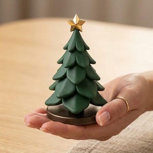 Peut inclure: Une petite figurine de sapin de Noël vert foncé stylisée avec des branches en couches en forme de pétales et une étoile dorée métallique au sommet. L'arbre est monté sur une base circulaire de couleur bronze foncé, formant un ornement compact.