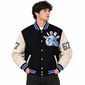 Puede incluir: Una chaqueta universitaria negra con mangas de color crema, que presenta un gráfico de casco de fútbol americano en azul y plata con el texto LIONS encima. Las mangas muestran los números 7 y 67, y el cuello y los puños tienen rayas azules y blancas.