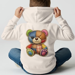 Può includere: Una felpa con cappuccio color crema con un colorato disegno di orsetto patchwork sul retro. L'orso ha toppe blu, marroni, rosse, verdi e rosa. Il bambino indossa jeans blu e si tira su il cappuccio.