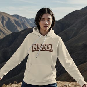 Może przedstawiać: Kremowa bluza z kapturem z napisem "MAMA" wyszytym na piersi brązową i białą czcionką w stylu uniwersyteckim. Bluza z kapturem ma kaptur ze sznurkiem i przednią kieszeń. Tło przedstawia pasmo górskie.