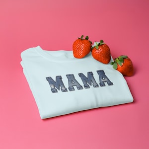 Może przedstawiać: Jasnoniebieska koszulka z napisem "MAMA" w stylu dżinsowym. Trzy świeże truskawki spoczywają na koszulce na różowym tle. Koszulka jest starannie złożona.