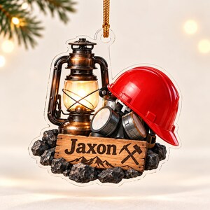 May include: A decorative hanging ornament featuring a bronze-colored lantern, a bright red hard hat, and a black respirator mask resting on a pile of coal. A wooden sign in the foreground displays the name Jaxon and a crossed hammer icon.