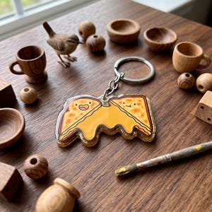 May include: A whimsical keychain featuring a smiling grilled cheese sandwich design with gold glitter accents. The image also includes wooden decorative items like a bird figurine, small bowls, beads, and a paintbrush, all arranged on a wooden surface.