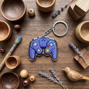 May include: A blue video game controller keychain with a silver keyring. The controller has a cat-like design with colorful buttons. The background includes wooden bowls, beads, a paintbrush, and a wooden bird on a wooden surface.