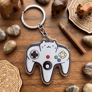 May include: A clear acrylic keychain featuring a white cat-shaped controller design. The controller has a black directional pad, a red button, and colorful buttons. It is attached to a silver keyring.