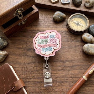 May include: A pink and white badge reel with the text "Hold on, Let Me Overthink This" in a speech bubble design. The badge reel has a clear plastic clip and a retractable cord. The background includes a wooden box, a compass, and decorative stones.