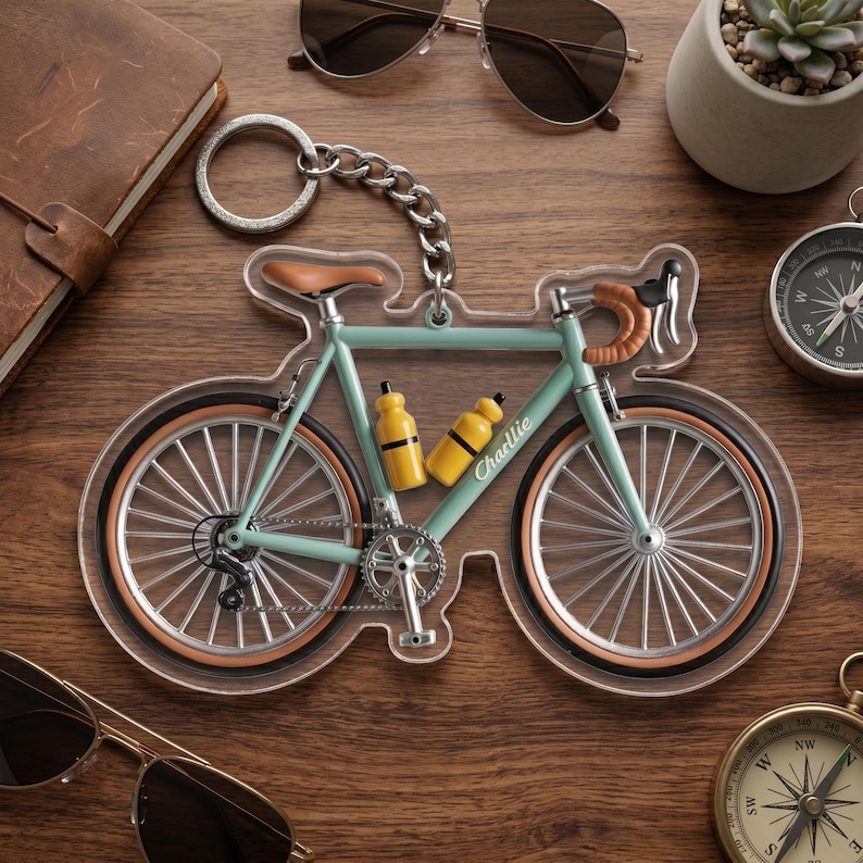May include: A bicycle-shaped keychain with a light blue frame, brown seat and handlebars, and two yellow water bottles. The word "Charlie" is printed on one of the bottles. The keychain is on a wooden surface with sunglasses and a compass.