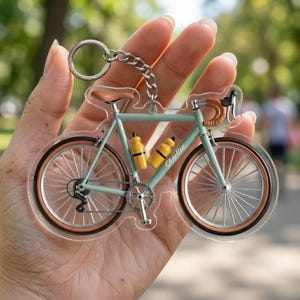 May include: A clear acrylic bicycle keychain with a silver chain and ring. The bicycle is light blue with brown accents, yellow water bottles, and the name "Charlie" printed on the frame. The wheels have a brown rim.