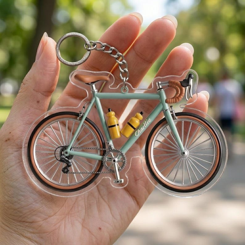 May include: A clear acrylic bicycle keychain with a silver chain and ring. The bicycle is light blue with brown accents, yellow water bottles, and the name "Charlie" printed on the frame. The wheels have a brown rim.