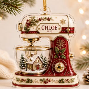 May include: A clear acrylic Christmas ornament shaped like a stand mixer, featuring a red and cream color scheme with gold accents. The mixing bowl contains a miniature snowy house scene with pine trees. The text CHLOE 2026 is printed on the front.