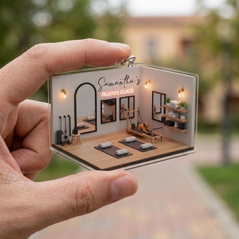May include: A clear acrylic keychain featuring a miniature Pilates studio. The keychain includes a Pilates reformer, mats, mirrors, and the text "Samantha's Pilates Class". The studio is decorated with small lights and shelves.