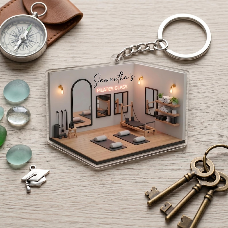 May include: A clear acrylic keychain featuring a miniature Pilates studio scene. The keychain has a silver chain and ring. The studio has a Pilates reformer, mats, mirrors, and the text "Samantha's Pilates Class".
