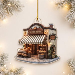 May include: A flat, shaped acrylic ornament depicting a bakery storefront with a brown and white striped awning, a wooden sign reading Chloe's Bakery, and shelves filled with various baked goods, suspended by a thin gold cord for hanging.