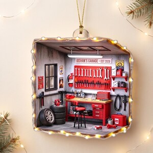 May include: A square hanging ornament depicting a miniature garage scene with a red workbench, tool pegboard, and a stack of tires. The sign on the wall reads JOSHAN'S GARAGE - EST 2020. The structure is outlined with warm white fairy lights.